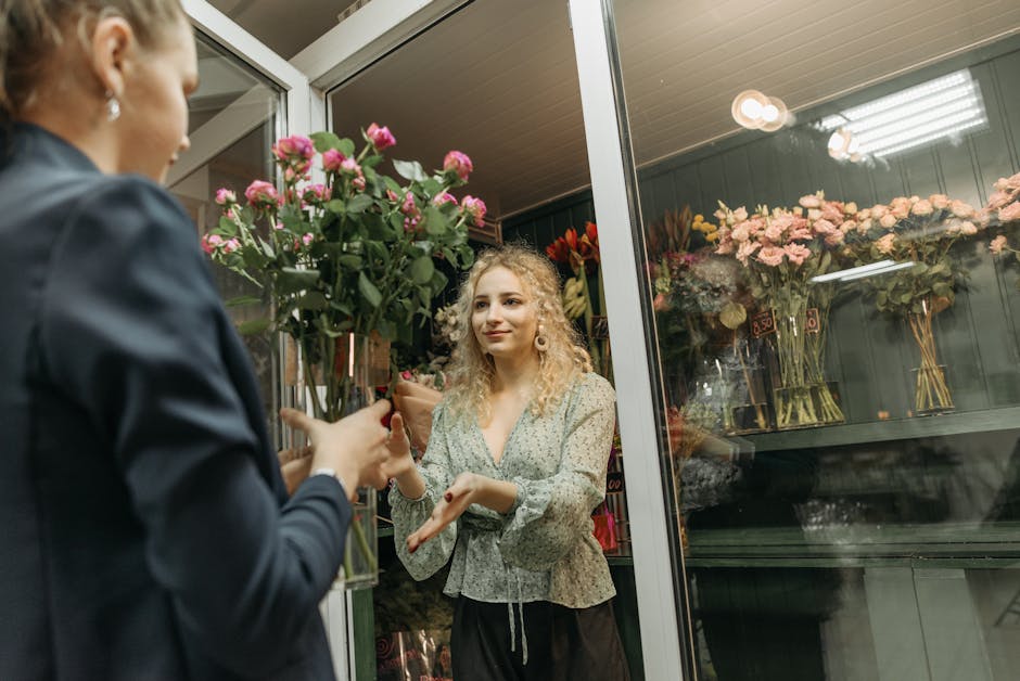 A young woman with curly blonde hair and fair skin, wearing a light-colored floral blouse, is engaging in a floral gift exchange at a flower shop. She is receiving a bouquet of pink roses with green f