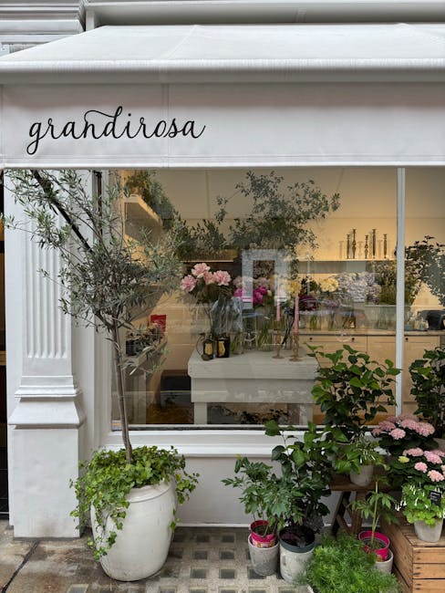 View of a flower shop exterior labeled 'Grandirose' with a large white canopy overhead. In front of the shop, there are potted plants, including an olive tree in a tall white pot, and several other gr