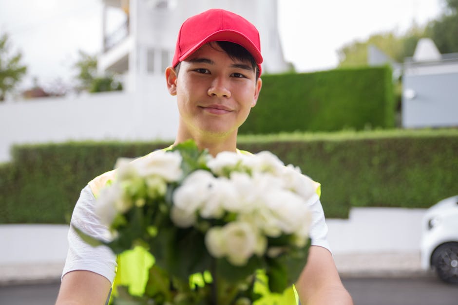 A friendly delivery person wearing a bright yellow vest and a red cap presents a bouquet of fresh white flowers, including roses and lilies, wrapped in clear cellophane with green accents. The bouquet