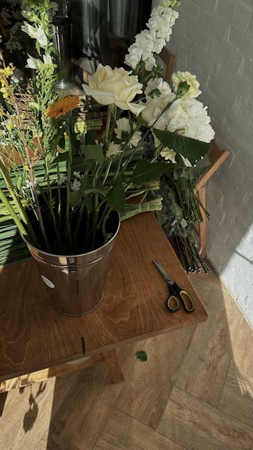 A tall, silver metal bucket filled with a fresh floral arrangement placed on a wooden table. The bouquet includes large, creamy white roses and white stock or delphinium with tall, spiked stems, compl