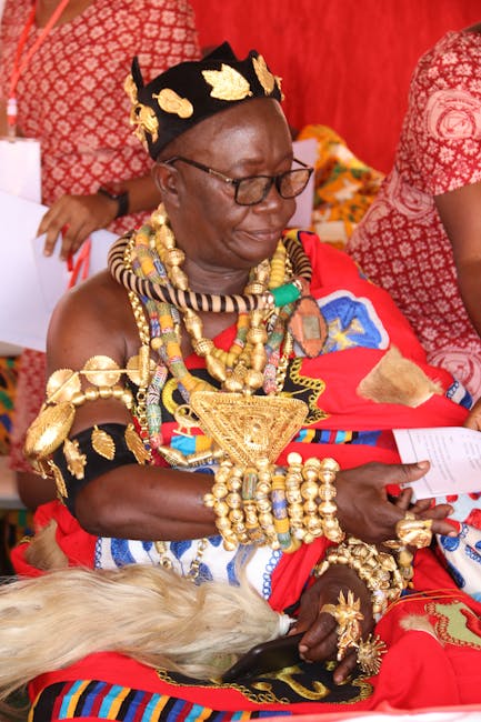 The image features an elderly woman with dark skin, wearing traditional African attire adorned with numerous gold jewelry pieces including necklaces, bracelets, and arm bands. She has a black headscar