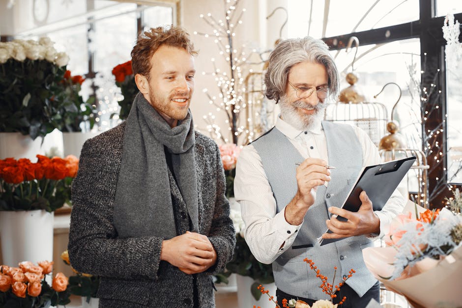 A male customer and a florist inside a flower shop engaged in a discussion about floral arrangements. The customer, with light brown hair and a beard, is wearing a dark tweed coat and a gray scarf, ho