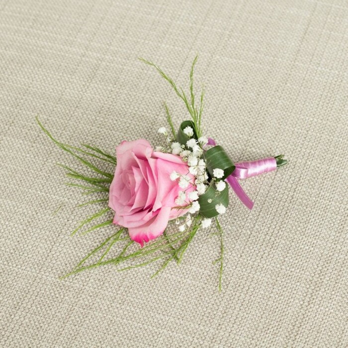 A delicately composed pink rose boutonniere lies on a light beige, linen-like fabric, whose fine, woven texture forms a calm and natural backdrop. The image is taken from a slightly overhead perspective, so you can appreciate the careful balance of each floral element. At the heart of the design is a single semi-open rose in blush pink tones, its petals layered tightly at the core and gradually loosening toward the outer edges. The colour transitions gracefully from a soft, warm blush at the centre to a deeper, more pronounced pink along the edges of the velvety petals, conveying both romance and freshness. Nestled snugly around the base of the rose, clusters of white baby's breath appear like miniature, airy clouds, their countless tiny flowers offering a gentle, textural contrast to the smooth rose petals. Emerging from behind the central bloom are slim, grass-like blades of bright green foliage that fan outward in fine arcs, giving the boutonniere a sense of movement and lightness. A slightly broader, darker green leaf is folded and wrapped around the lower portion of the stems, functioning as a natural collar that neatens and supports the compact design. The stems are meticulously bound with a satin ribbon combining soft lavender-pink with a richer magenta-purple sheen, tied securely at the base in a simple knot that leaves the focus on the flowers. Soft, even lighting casts faint, flattering shadows, enhancing the three-dimensional form without distracting glare. The overall impression is of a timeless, handcrafted accessory perfectly suited to a groom's lapel for a ceremony in Gerrards Cross, perhaps at a venue near West Common, or for a prom date meeting under the trees by the local green, carrying with it a quiet air of celebration and refined style.