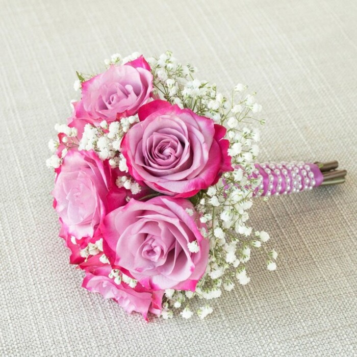 This elegantly composed wedding bouquet, reminiscent of a scene from a Gerrards Cross garden, features five exquisite roses whose petals display a seamless transition from deep, sumptuous magenta to delicate, pastel lavender towards the center. Interwoven are tiny clouds of white baby's breath-each floret handcrafted to float delicately among the lush, full blooms, giving the arrangement a romantic and gentle volume. Bound together, the short stems are meticulously wrapped in a rich plum satin ribbon, which is itself adorned with spiraling strands of lustrous white pearls, reminiscent of classic bridal accessories treasured in Gerrards Cross's storied wedding traditions. The bouquet's natural stems emerge at the base, lending an authentic, freshly gathered mood. It sits gracefully on a pale, textured linen cloth, the neutral hue providing a perfect backdrop for the bouquet's vivid florals and intricate handle. Captured from above, the round, harmonious shape is revealed, with each floral detail displayed clearly-a testament to careful craftsmanship and local artistry. Ideal as a bridesmaid's bouquet, for a flower girl walking the aisles of a historic Gerrards Cross church, or as a romantic focal piece for a spring wedding, this arrangement invites joyful celebration and timeless elegance to any special occasion.