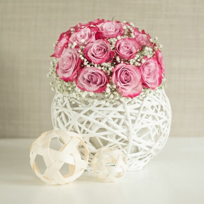 An elegant tabletop display features a dome of pink roses and white baby's breath standing proudly in a spherical white woven vase, with two light wooden decorative balls placed artfully to one side. At the centre of the composition, the bouquet is dense and rounded, built from numerous medium-sized roses in soft shades of pink. Each rose shows a gentle gradient of colour: the inner petals are a delicate, almost porcelain blush, while the outer petals deepen into a more saturated rose pink, some nearing fuchsia along their ruffled edges. Nestled between the roses are many sprays of gypsophila, their tiny white flowers and fine stems creating a misty, cloud-like halo that breaks up the solid mass of pink and introduces a feeling of air and movement. The flowers are arranged in a striking white vase formed from interwoven strands that criss-cross to create a hollow, globe-shaped structure, suggesting a modern take on wicker or rattan. The openwork design lets the eye glimpse through the vase, emphasising its sculptural quality. On the glossy white surface in front of it rest two decorative spheres, each made from thin, beige wooden strips woven into open, geometric forms, their natural tone adding warmth and a hint of Scandinavian-inspired simplicity. Behind, a softly textured wall in a warm off-white or light beige offers a quiet backdrop, similar to the calming interiors found in many Gerrards Cross homes just off Oxford Road, and helps the pinks and whites appear fresh and luminous. The lighting is bright yet diffused, with no harsh shadows, enhancing the textures of petals, wicker and wood. The overall feeling is one of serene sophistication and gentle romance, ideal for marking anniversaries, housewarmings or simple thank-you gestures, especially when imagined as a fresh delivery arriving straight to a doorstep in Gerrards Cross.