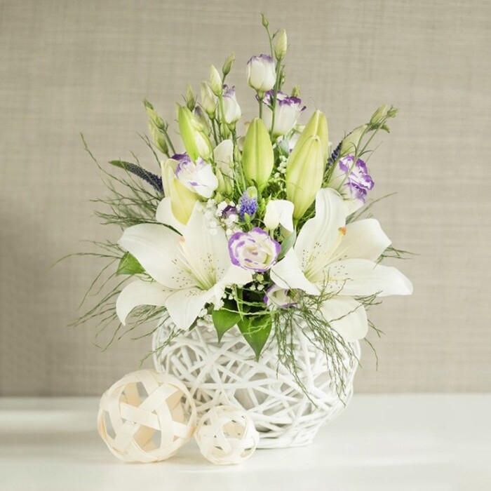 In this refined floral composition, a rounded white lattice vase holds a graceful mix of white and lilac-hued blooms, styled much like a wedding table arrangement ready for a reception in Gerrards Cross. Several tall stems of white lilies dominate the arrangement, some fully opened into wide, trumpet-shaped flowers showing fine veins and soft shadows within the petals, others still enclosed as sleek, light green buds that curve delicately upwards. Interlaced with them are white lisianthus (eustoma), their blooms layered and ruffled, with many petals edged in pale lavender or washed with soft purple at the throat, lending a romantic, almost bridal quality. Smaller clusters of deep purple filler flowers peek through like little jewels, while airy white sprays, similar to baby's breath, fill the gaps and lend a cloud-like softness. Around and beneath the blooms, a generous collar of mixed foliage creates a lush framework: feathery green sprigs, slender looping stems, and slightly broader, more structured leaves spill naturally over the vase edge, as if freshly gathered from gardens near Bulstrode Park. The vase itself is striking, crafted in a spherical shape with an open-weave pattern reminiscent of rattan or wicker, painted a crisp white that harmonises with the petals and echoes contemporary interior trends. On the smooth white tabletop, two decorative woven balls in light beige and natural tones sit to one side, repeating the textured motif and balancing the visual weight. The softly lit background is a light beige wall with a gentle horizontal, linen-like texture, catching the light just enough to frame the arrangement without distraction. The overall impression is serene, pure, and slightly ceremonial, ideal for a wedding table, a remembrance service, or a calm focal point in a Gerrards Cross living or dining room.