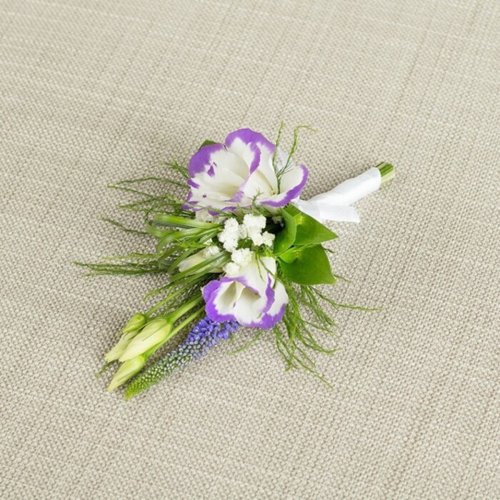 This image shows a finely detailed floral boutonniere arranged on a softly textured, light beige fabric that resembles natural linen, the neutral shade providing a calm, elegant backdrop. At the centre are two striking lisianthus blooms: their petals a clean, luminous white, each edge deeply ruffled and delicately tipped with saturated purple, creating a vivid bi-colour effect that feels both romantic and modern. Around these focal flowers sit dainty sprays of small white filler blooms, similar to baby's breath, which add a sense of lightness and gentle movement, as though a soft breeze has just passed over a garden on the quiet streets near West Common in Gerrards Cross. A slim, vertical stem of tightly clustered, deep purple, bell-shaped flowers-resembling muscari or grape hyacinth-threads upward beside the lisianthus, introducing a more intense shade of purple and an interesting, bead-like texture. Fine, feathery greenery, akin to asparagus fern, fans out around the blooms, its delicacy echoing the soft touch of moss or light foliage underhand, while a few slightly broader green leaves provide structural balance and depth. Two pale green buds are included in the arrangement, their closed forms hinting at blossoms still to come and symbolising new chapters beginning, a fitting sentiment for weddings or milestone celebrations. The stems of all the flowers and foliage are gathered neatly and wrapped in a smooth white satin ribbon, secured with a small, tidy bow that sits flush against the stems, clearly indicating that the piece is ready for pinning to a lapel without fuss. Prepared with the kind of care a local florist brings to Garrards Cross wedding parties, this boutonniere feels bespoke and personal-ideal for a groom, groomsman, or formal guest wanting a refined, understated accent. The overall mood is sophisticated yet approachable, harmonising classic white and green with rich purple tones for a timeless, photograph-friendly finish.
