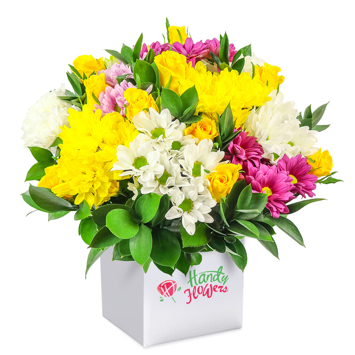 A vibrant mixed bouquet bursts from a pristine white square box, captured against a seamless white background that enhances its lively colours and structured shape. The arrangement is tightly composed into a generous dome, full and overflowing yet carefully balanced. At its heart are large yellow chrysanthemums, their petals densely layered to form soft, plush cushions of colour reminiscent of late-summer sunshine over Gerrards Cross Common. Around them, clusters of yellow spray roses appear in varying stages of bloom, from small rounded buds to partially open flowers, offering an engaging mix of textures and a sense of natural growth. Fresh white daisy-like chrysanthemums with green centres create a bright ring of contrast, while plumper white chrysanthemums add airy volume and a gentle, cloud-like softness. Woven artfully through these lighter tones are vivid fuchsia and magenta chrysanthemums, some with touches of pale pink at the edges, providing deep, jewel-like notes that give the bouquet depth and sophistication. Small pink and yellow rosebuds are tucked here and there, almost like hidden treasures, lending a romantic, thoughtful detail. The base of the design is surrounded by lush, deep green foliage, likely ruscus or a similar sturdy leaf, its glossy surfaces framing the blooms and anchoring the arrangement. The smooth white box functions as both container and presentation gift, with clean lines that suit modern homes around Gerrards Cross, from new builds near the train station to period houses off Oxford Road. The overall effect is cheerful yet refined, ideal for birthdays, congratulations, or simply brightening a room with the feeling of fresh flowers just delivered by a local florist who knows the area well.