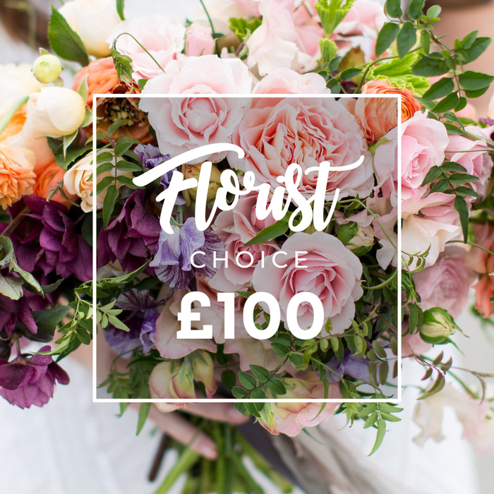 A sumptuous, florist-selected bouquet sits at the centre of the photograph, its layered textures and hues framed by a narrow white rectangle bearing the words Florist Choice in flowing script, CHOICE in smaller block letters below, and a large, bold ?100 beneath - an elegant label that tells you this is a premium, florist-chosen arrangement. The bouquet reads like a romantic garden composition: multiple large pink garden roses with ruffled, velvety petals take centre stage, their soft blush tones contrasted by warm peach and coral ranunculus that offer luminous, candy-coloured depth. Darker, plum-hued hellebore-like flowers cluster toward the lower left, grounding the palette with velvety richness, while pale purple-and-white striped sweet peas and small creamy-white filler blooms breathe lightness between the larger heads. A generous selection of greenery - from slender, trailing vines to broader, sculptural leaves - is woven throughout to create movement, fullness and a freshly gathered silhouette. The stems are gently held at the base by human hands; a faint white sleeve and blurred background keep the bouquet intimate and tactile, as if just picked and tied in a shop near Gerrards Cross High Street. The soft daylight highlights petal layers and the subtle scent one imagines from the image; the overall effect is handcrafted luxury and seasonal abundance. This photograph communicates careful florist craftsmanship, a thoughtful gift choice for anniversaries, weddings or an indulgent, surprise delivery across Gerrards Cross and neighbouring villages.