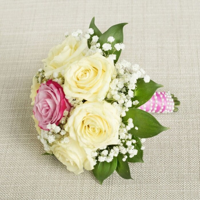 Presented atop a softly woven, light beige linen reminiscent of the welcoming interiors found across Gerrards Cross, this dainty bouquet is a study in refined celebration. A cluster of fresh, creamy roses dominates the compact arrangement, their petals pristine and gently unfurling at the centre, hinting at both innocence and joy. Set just to the left, a striking magenta-pink rose captures attention, infusing the delicate palette with a sense of bold romance perfectly suited for wedding festivities in the tranquil neighborhoods of Gerrards Cross. Scattered amidst the roses are clouds of white baby's breath, their tiny blossoms lending a gentle, airy texture and an enduring sense of bridal tradition. Surrounding the flowers, supple dark green leaves provide contrast and enhance the impression of natural abundance. The stems are elegantly wrapped in a polished, fuchsia-pink satin ribbon, meticulously finished with spirals of soft white pearls that gleam subtly in the late morning light. Each element, from petal to pearl, has been arranged with thoughtful expertise by local florists who understand the timeless appeal of understated elegance. The bouquet-and the gentle, fresh fragrance it suggests-evokes the promise and romance of special occasions across Gerrards Cross, from a chic registry office gathering to a private celebration in one of the area's leafy gardens. This image encapsulates the warmth and artistry of a bouquet designed for unforgettable moments.