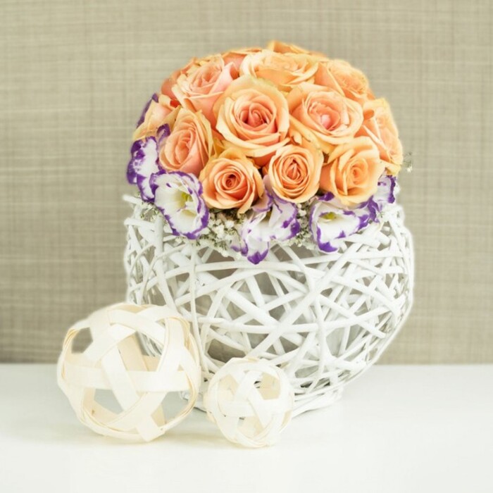 On a pristine white tabletop, a carefully composed floral centerpiece radiates gentle romance, the sort of arrangement that might grace a wedding breakfast at a Gerrards Cross hotel or a stylish family gathering near Bulstrode Park. The focal point is a dense, rounded cluster of roses in soft peach and apricot shades, each flower fully open and nestled closely to its neighbours, so that the rose heads merge into a smooth, cushioned dome of petals. Encircling the base of this rose sphere is a halo of white lisianthus blooms, their frilled petals delicately outlined in deep violet and purple, adding a striking yet elegant contrast. Interspersed among them are airy stems of white gypsophila, which drift like tiny clouds between the larger blossoms and bring a light, feathery texture to the design. The arrangement is held in a distinctive white container shaped like a broad, rounded bulb, meticulously woven from rattan or wicker into an open lattice pattern that lets the background gently show through. In the foreground on the same shining surface rest two decorative orbs crafted from pale, natural wood-like strips, one larger and one smaller, their open-weave construction echoing the basket's design and creating a cohesive, textural story. Behind the flowers, a subtly textured beige fabric backdrop-suggestive of soft linen-provides a warm, neutral setting that allows the peach, white, and purple tones to glow without distraction. This image conveys a mood of natural elegance and thoughtful craftsmanship, ideal for illustrating wedding table arrangements, refined dinner party décor, or special occasion flowers delivered throughout Gerrards Cross.