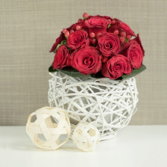 On a smooth white table, a rounded bouquet of crimson roses forms a perfect, compact dome, ideal as a wedding table centrepiece for a celebration in Gerrards Cross. The roses are rich and velvety, their petals tightly layered so that each bloom looks plump and luxurious, with subtle variations in tone from deep wine red to slightly lighter scarlet at the tips. Nestled shyly between the blooms are clusters of small red hypericum berries, adding gentle pops of shine and an inviting, berry-like texture that contrasts with the softness of the petals. Just beneath the lush crown of flowers, a neat collar of dark green leaves peeks out, offering a hint of foliage that frames the arrangement without disrupting its sculptural, almost spherical form. The entire bouquet rests in a unique, openwork vase crafted from white-painted twigs or slender branches, woven into a hollow globe with plenty of negative space. This light, airy structure allows glimpses of the green stems inside and creates subtle patterns of shadow where the twigs cross one another. The bright, even lighting gives the whites a crisp clarity and makes the reds appear especially saturated, as though freshly arranged by a local florist ready for same-day delivery across Gerrards Cross and nearby areas. To the left, on the same white surface, sit two decorative wicker balls in natural, pale wood tones, one medium-sized and one smaller, echoing the open, lattice design of the vase and providing a soft, organic counterpoint. In the background, a neutral wall in light beige or taupe, perhaps reminiscent of a calm dining room near West Common, is softly blurred, ensuring that the rich colour and texture of the roses remain the undisputed focal point.