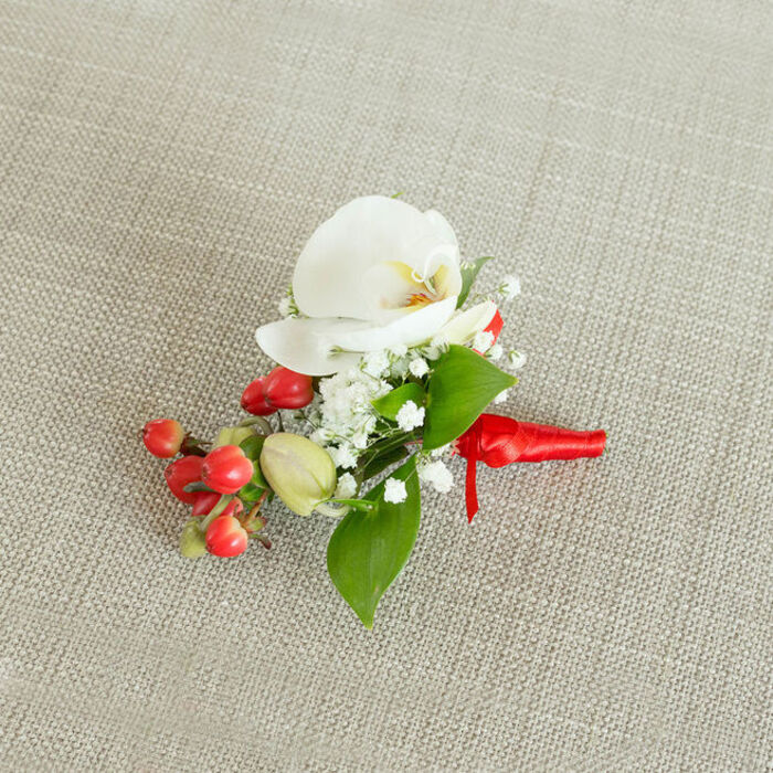 An artfully composed boutonniere lies against a softly textured, oatmeal-coloured linen background, the neutral fabric allowing every floral detail to stand out with clarity and grace. At its centre is a luminous white Phalaenopsis orchid, the petals wide and gently curved, forming an open, sculptural shape that instantly suggests elegance and calm. The orchid's throat holds a delicate blend of creamy yellow and soft orange, like a warm glow at the heart of the flower, adding depth and a hint of warmth to the otherwise cool, white palette. Surrounding this focal bloom are airy sprays of baby's breath, their countless tiny white florets forming a cloud-like border that softens the lines of the orchid and creates a sense of lightness. Interwoven amongst the flowers are smooth green leaves in varied forms-some broad and pointed, others more slender and elongated-placing the bloom firmly in a natural, garden-inspired context, as if freshly gathered from a well-tended plot not far from the green spaces of Gerrards Cross. On one side of the boutonniere, a cluster of glossy red berries draws the eye, their rich, saturated colour echoing the deep reds often seen in winter weddings or festive celebrations. A few lighter green, berry-like buds offer a subtle contrast, suggesting new life and the promise of the season ahead. The stems are carefully bound with a vibrant red satin ribbon, wrapped smoothly and secured in a small, tidy knot with short tails that lie flat and controlled. The overall composition feels deliberately balanced and thoughtfully made, the kind of refined floral accessory chosen for a groom, best man, or guest attending a special celebration near the centre of Gerrards Cross, where attention to detail and understated luxury are highly valued.