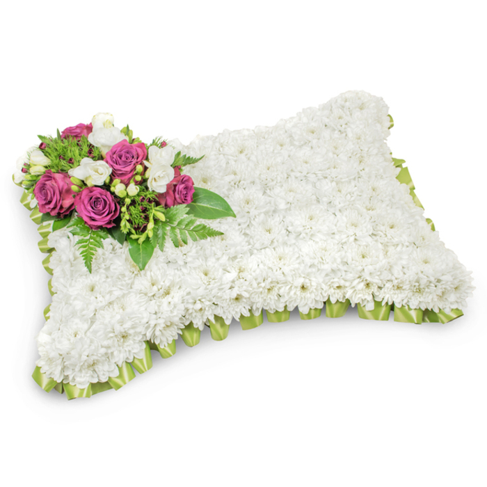 A rectangular sympathy pillow, meticulously assembled from hundreds of tiny white chrysanthemum blooms, presents as a soft, uniform expanse that evokes comfort and serene repose. Each chrysanthemum is placed to create a seamless, tactile surface-rounded at the corners and plush to the eye-so the arrangement reads like a true cushion of flowers. In the upper-left quadrant a composed spray of deep magenta roses becomes the arrangement's heartfelt accent: their velvety petals open in rich layers, surrounded by fragrant white freesias and small white buds that lift the composition with delicate highlights and a faint floral perfume. Beneath and around these focal blooms, graceful fern fronds and glossy greenery fan out, offering a fresh, supportive base and a cool contrast to the warm roses. The rim of the pillow is finished with a pleated light-green satin ribbon that gently peeks out beneath the chrysanthemums; its subtle shine and neat loops give the piece a thoughtful, ceremonial edge. Photographed against a crisp white backdrop with a soft shadow to define its three-dimensional form, this classic design is intended as a dignified tribute for funerals, wakes or graveside services in Gerrards Cross and surrounding communities such as Burnham and Denham Country Park. Prepared with the care you'd expect from a local florist, this arrangement-available through Flower Delivery Gerrards Cross-offers solace through texture, colour and quiet craftsmanship when families gather to remember.