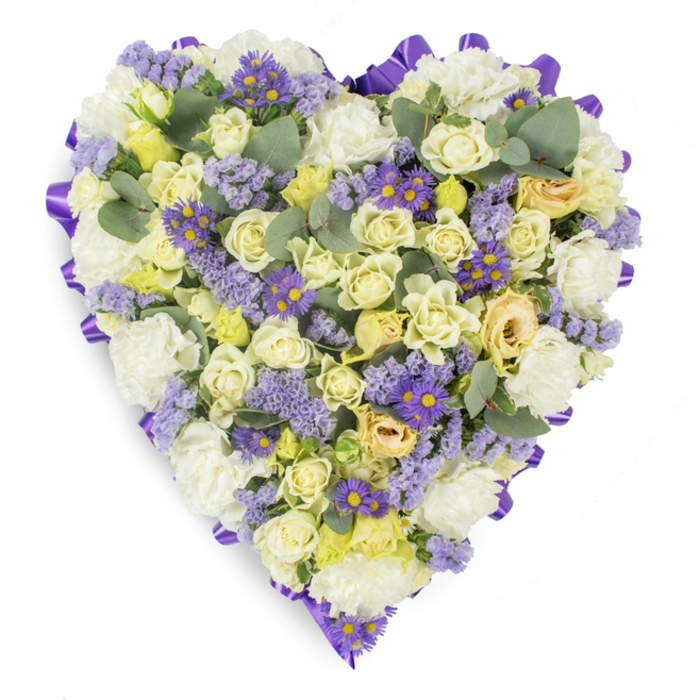 A refined heart-shaped sympathy arrangement shown against a pristine white backdrop, composed with close-set blooms to form a compact, contemplative surface. The dominant palette is soft and restrained: creamy roses in shades of pale yellow and ivory create a plush, tactile center, their petals ranging from tight, rounded buds to gently unfurling blossoms that add depth and subtle variation. Interwoven with these are abundant white carnations whose frilled edges give a comforting, classic texture. Cooler lilac elements - sprigs of statice and small purple, daisy-like aster flowers with conspicuous yellow centers - are distributed across the composition to introduce delicate contrast and a measured brightness without disturbing the overall calm. Blue-green eucalyptus leaves, rounded and slightly glaucous, punctuate the massing of blooms, their shapes and tones providing elegant breaks in the density and a suggestion of fresh, herbal fragrance. The heart's edge is consciously defined by a neatly folded purple satin ribbon running continuously around the perimeter, giving a finished, ceremonial frame suited to memorial use. The lighting is even and soft, accentuating petal surfaces and foliage sheen while maintaining a subdued, respectful mood. This type of tribute is well suited to funerals, memorial services and graveside remembrances in Gerrards Cross and surrounding parishes, offering an expression of enduring affection, solace and dignity.