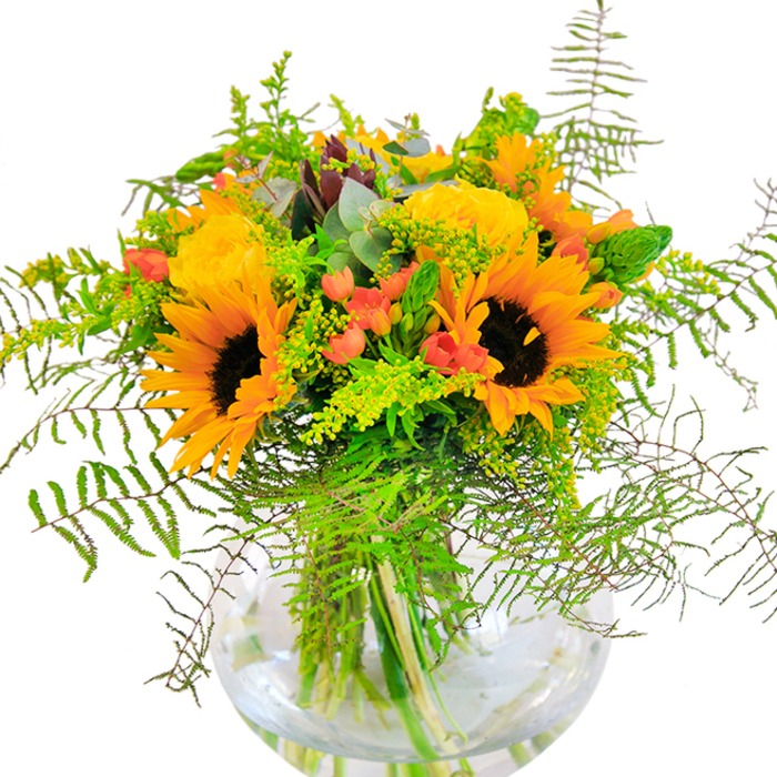 At first glance, the room is awash in a cheerful palette of golden yellows and rich greens, centered by a bouquet that thrives on sunlight and spirited abundance. Prominent sunflowers claim attention, their radiant petals and bold, textured centers drawing the eye as naturally as a stroll towards the shops of Gerrards Cross on a bright day. Intertwining with them, buttery garden roses spiral in intricate rosettes, their fullness imparting a sense of luxury and warmth. Ruby-like clusters of Hypericum berries peek out with playful energy, providing a striking visual rhythm among the vegetation. Delicate fronds and wisps of green-reminiscent of the wild fern edges along East Common-spill gently over the rim, lending the arrangement a sophisticated sense of movement and grace. Subtle accents of blue-green eucalyptus leaves recall the freshness of a dewy morning in the local woodlands, their unique scent wafting softly. Anchoring this lively display is a crystal-clear, round glass vase: its see-through walls reveal the ordered, verdant stems submerged in fresh water. This composition is perfect for commemorating milestones, welcoming friends, or enriching everyday moments in and around Gerrards Cross. Every petal, leaf, and berry whispers of local meadows, capturing the season's warmth and inviting celebration in any space.