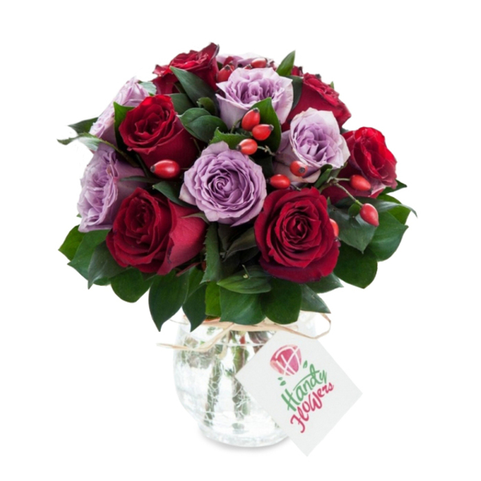 An expertly composed bouquet that marries deep, sensual reds with soft, cool lavenders: velvet crimson and burgundy roses are interlaced with lilac and dusty purple roses, each bloom fully open and revealing layers of petal that catch the light like silk. Rich, dark green foliage - likely Ruscus or Salal - forms a structured underlayer, its glossy leaves creating negative space that allows the rose colours to sing without overcrowding. Bright red-orange rose hips or small berry clusters are artfully tucked into the design, bringing sparks of warm colour and a firmer texture that contrasts with the roses' satin softness. The flowers are arranged in a rounded silhouette and presented in a rounded mercury-glass vase with a mottled, antique silver sheen; the vase's reflective surface skews highlights and shadows, so the bouquet reads as both elegant and slightly rustic. A natural raffia or twine is tied around the vase's neck and supports a small white rectangular gift tag - blank here of branding - ready for a handwritten note. The whole scene is shot on a crisp white background that accentuates the intensity of the reds and the delicate cool of the lavenders, and it conveys an atmosphere of romance, considered gifting and premium floristry. As a touch of local provenance, this is the sort of arrangement our team at Flower Delivery Gerrards Cross might prepare for an anniversary delivery to a home on the High Street or a celebratory bouquet collected from the shop near Beaconsfield Road.