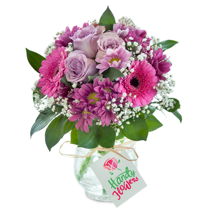 Delicately poised on a crisp white surface, this exquisite floral arrangement captures the fresh, welcoming spirit of Gerrards Cross in a vivid celebration of color and form. The composition begins with a cluster of pale lavender roses, each blossom soft and gently expanding, lending an air of romance and tranquility. Radiating outward are eye-catching hot pink Gerbera daisies, their broad petals and inky centers creating dynamic contrast and cheerful energy-much like the vibrant corners of Windsor Road in bloom each spring. Interspersed throughout, deep magenta and plum-toned chrysanthemums provide intriguing visual layers, while sprigs of snow-white baby's breath elevate the bouquet with a light, ethereal touch. Lush, glossy emerald leaves peek from beneath the blooms, grounding the creation with a natural flair reminiscent of local woodlands. All are artfully arranged in a clear glass vase with a distinctive 'cracked ice' pattern, further enhanced by a loop of rustic raffia tied at its neck and a minimal white tag ready for a hand-written note. Crafted with an expert's care and deep local knowledge, this vibrant bouquet stands as a testament to Gerrards Cross floristry-an ideal gift for any special moment or to bring a touch of blooming delight into daily life, all available for delivery just moments from the station.