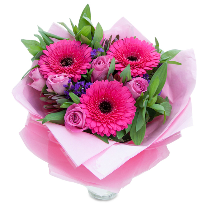 There is a particular charm in pinks that lean toward both playful and refined, and this bouquet captures that balance in a tactile, expressive way. Three fuchsia Gerbera daisies command attention with their broad, perfectly formed petals and dark centres that provide a striking graphic contrast. Around them, roses in a dusty lavender-pink remain gently furled-those early-stage petals hold scent and promise, a soft perfume of tea-rose that fills a small room without overwhelming it. Intermittent sprays of purple statice lend a wildflower note and a change in texture-tiny clustered blooms that seem to float between the larger faces of Gerbera and rose. The greenery is generous and purposeful, with glossy leaves resembling salal and slinky eucalyptus stems that add a cool, slightly resinous aroma and visual depth. The wrapping is part of the design: an inner sheet of matte pale-pink tissue cushions the stems, while an outer sash of translucent fuchsia-pink cellophane, faintly crinkled, adds shimmer and shapes the silhouette. You can see the top rim of a clear glass vase peeking at the bottom edge, an invitation to set this arrangement out straight away. As someone who arranges flowers for neighbours from Gerrards Cross Station to the village green, I imagine this placed on a breakfast table after a walk in Black Park or Burnham Beeches-bright, reassuring, fit for birthdays, thank-you gestures, or simply to brighten the afternoon light.