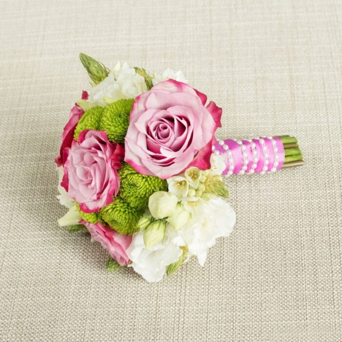 Arranged horizontally on a softly textured, light beige fabric, this small bouquet feels like a captured moment from a quiet wedding morning in Gerrards Cross. The fabric surface beneath, reminiscent of fine linen or burlap, has a gentle weave that adds a rustic, natural backdrop without competing with the flowers. Centred in the design, three pink roses form the main focal point, with two especially prominent: one at the upper centre and one slightly lower and to the left. Their petals unfurl in generous layers, satin-smooth and delicately curled at the edges. The colouring moves from a pale, almost shell-pink at the core to a deeper rose and finally to rich, magenta-tinged edges on the outer petals, giving each bloom depth and a sense of hand-painted detail. Tucked snugly among the roses are clusters of lime-green button chrysanthemums, their tiny, tightly packed florets forming perfect spheres. These green buttons add a lively pop of colour and a pleasing, almost tactile texture that invites the fingertips to explore, like mossy cushions in a shaded garden near East Common. Scattered throughout the bouquet are smaller blossoms in tones of white and cream-delicate filler flowers that might be lisianthus, freesia, or wax flower-some with tiny, star-shaped petals and faint yellow centres. They weave through the arrangement, creating a lace-like lightness around the more substantial blooms. Slender hints of darker green foliage peek through, linking everything back to a natural, garden-grown origin. The stems, trimmed short and even, are wrapped tightly to form a neat handle. Over them, a vibrant fuchsia satin ribbon is wound with care, its surface catching highlights in the soft, even lighting. A slender strand of white faux pearls loops diagonally across the ribbon, as if a favourite necklace has been repurposed to adorn the bouquet, adding a touch of timeless, bridal elegance. The overall composition feels intimate and refined, perfect for a bridesmaid, a petite bridal posy, or an elegant table arrangement for a Gerrards Cross celebration, radiating freshness, romance, and quiet sophistication.
