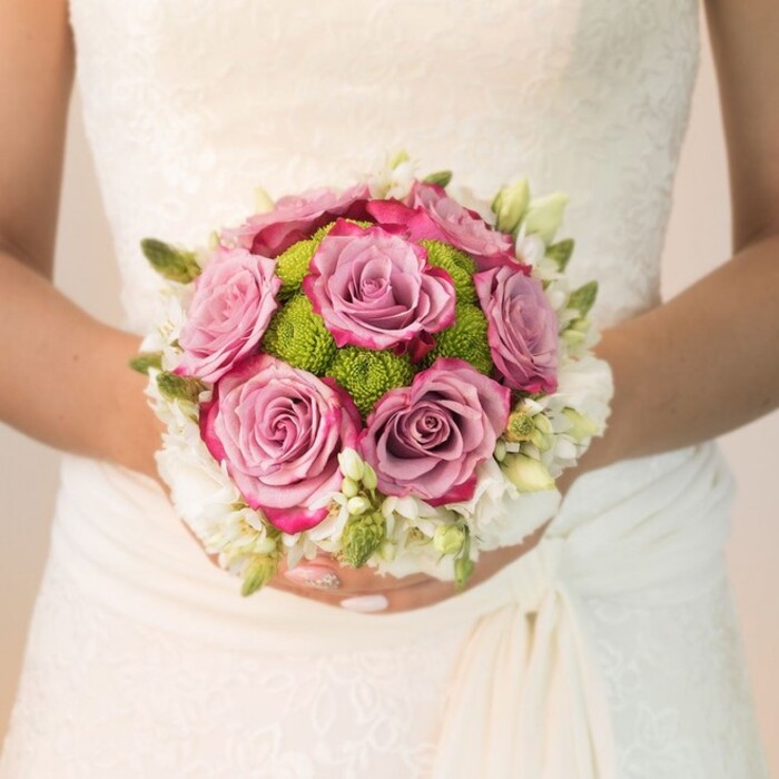 Captured at waist-level, this image details the refined beauty of a Gerrards Cross bridal bouquet, thoughtfully designed for a timeless wedding celebration. The bride, her hands delicately adorned with shimmering blush-toned nail polish, supports a compact, rounded arrangement that rests elegantly against the intricate lace and soft silk folds of her off-white gown-inspired perhaps by the tranquil elegance of events along Packhorse Road. At the bouquet's core, large roses bloom in shades of dusty mauve and luminous lavender-pink, their ruffled edges whispering hints of magenta and fuchsia for a dynamic, multi-layered effect. Surrounding the roses, a captivating circle of lime-green button chrysanthemums injects vibrant texture, echoing the bold foliage found in Gerrards Cross's private gardens and green spaces. The bouquet's edge is delicately framed with clusters of white blossoms, possibly freesia or miniature spray roses, accompanied by slender green buds and organic sprigs, imparting a light, romantic freshness. Subtle natural lighting washes warmly over both the gown's lace detail and the bouquet, highlighting every petal's softness and the organic complexity of the arrangement. The overall composition speaks to the sophisticated simplicity often chosen by brides in Gerrards Cross, uniting pastel and bright colors within a timeless design. Perfect for wedding features, floral inspiration, or discussions of local bridal style, this bouquet reflects the artistry and heartfelt care invested in every wedding flower order at Flower Delivery Gerrards Cross.