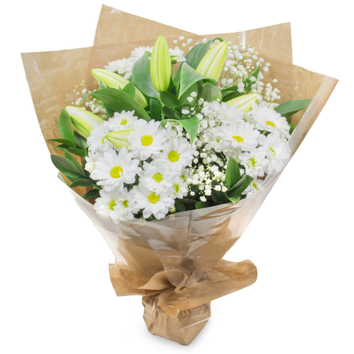 Presented upright on a stark white field, this bouquet is a study in calm contrast and gentle anticipation: clustered around the centre are numerous white chrysanthemum blooms-daisy-like, petal-rich, each with a small yellow eye-that create a soft, luminous core. Rising among them are several long, pale green-yellow lily buds, some bearing faint stripes, their poised, unopened shapes introducing a refined verticality and the suggestion of future bloom. Fine clouded branches of baby's breath (Gypsophila) are peppered throughout the arrangement, their tiny white florets giving an ethereal, almost drifting texture that mellows the stronger shapes. Broad, deep green foliage frames the flowers, the leaves' darker tones setting off the crispness of the whites and lending visual weight and depth. The bouquet is hand-gathered and wrapped in a tactile inner layer of crinkled brown kraft paper, which lends an earthy, artisanal feel, and an outer layer of translucent cellophane that glints softly in the light and keeps the composition pristine. The base is neatly folded and tied so the bouquet stands as a compact, portable piece, ready to be handed over or placed on a mantle. The overall aroma is delicate and green-fresh-cut stems and a faint, clean floral sweetness-evoking a morning market stall near Gerrards Cross High Street or a quiet delivery after a walk through Denham Country Park. This arrangement reads as pure and composed, suitable for marking moments from new beginnings and birthdays to expressions of sympathy, and reflects the care of an experienced local florist who values texture, restraint, and the gentle storytelling power of white blooms.