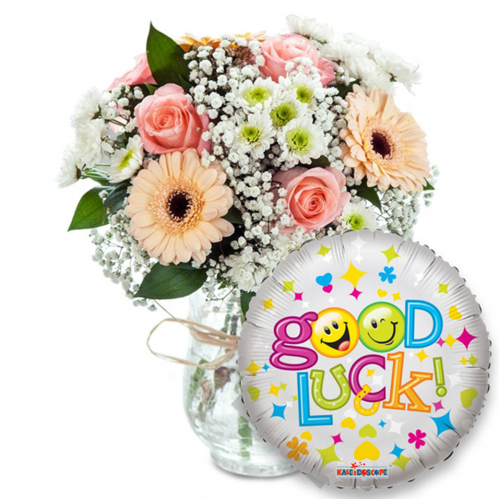 An encouraging gift ensemble shown against white: a lush bouquet fills the upper left and centre in a simple clear glass vase, its neck wrapped in rustic twine for an artisanal touch. The arrangement carefully balances soft peach roses and powder-pink roses-petals layered like silk-with sunny peach-orange Gerbera daisies whose deep brown centres add a strong visual counterpoint. Dainty clouds of white baby's breath (Gypsophila) weave throughout, lending a delicate, ethereal texture, while small clusters of white and lime-green chrysanthemum florets bring a clean, invigorating brightness. Dark green leaves tuck among the blooms, providing contrast and an earthy, leafy aroma beneath the floral perfume. In the foreground, to the right and slightly overlapping the vase, a round metallic silver foil balloon shines and reflects light; it's patterned with playful hearts, stars and tiny colourful shapes in pink, blue, yellow and green. Boldly across the balloon is the phrase Good Luck! in multi-coloured, bouncy lettering, and two of the round letter-forms are replaced by sunny yellow smiley faces-one winking, one sticking its tongue out-infusing the image with cheeky warmth. A small KALEIDOSCOPE mark appears at the balloon's base. The composition reads as a thoughtful send-off or celebratory token-perfect for graduations, farewells, or first-day nerves-and feels like something a local florist would hand over outside Gerrards Cross station or at a community gathering in the parks near Denham.