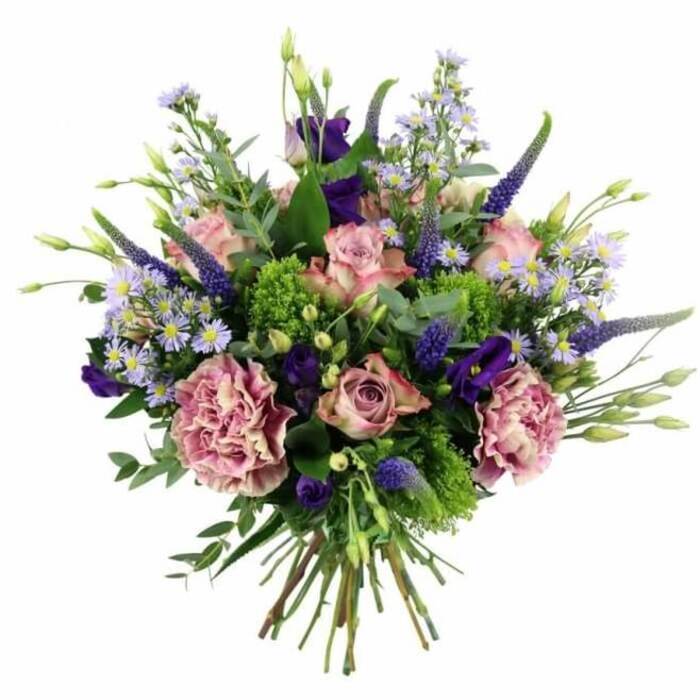 This image shows a beautifully composed hand-tied bouquet positioned against a bright, white backdrop, allowing every nuance of colour and texture to stand out clearly. The arrangement is dominated by a soothing palette of soft pinks, mauves, and purples, with roses taking pride of place at the centre. Some of these roses are still in bud, their petals wrapped tightly in a promise of full bloom, while others are more open, displaying layered whorls that shift gently from a deeper pink core to paler, almost creamy outer edges. Cradling and amplifying their romance are generously ruffled carnations in tones of dusty rose and purple-mauve, their intricate frills giving the bouquet a sense of depth and movement. From within this rounded cloud of petals, elegant spikes of Veronica arise, coloured in deep lavender-blue, lending both height and a slightly wild, garden-picked character that calls to mind the borders of gardens around Gerrards Cross station. Interwoven throughout the design are clusters of small, daisy-like, lavender and periwinkle blossoms, each with a delicate yellow centre, softening the bolder hues and adding a light, airy texture. Richer notes of purple come from lisianthus blooms, some cupped and some just beginning to open, bringing a velvety richness to the overall look. Surrounding all of the flowers is an abundance of foliage: rounded, silvery eucalyptus leaves, sprigs of vivid chartreuse-green filler, and other mixed greenery that keeps the bouquet feeling fresh and vibrant, like a walk across East Common after a spring shower. The stems are visible beneath the flowers, trimmed evenly and tied closely together, ready to slip into a favourite vase. The composition evokes warmth, love, and refined celebration, ideal for marking weddings, anniversaries, or milestone birthdays with a stylish flower delivery in Gerrards Cross and the nearby villages.