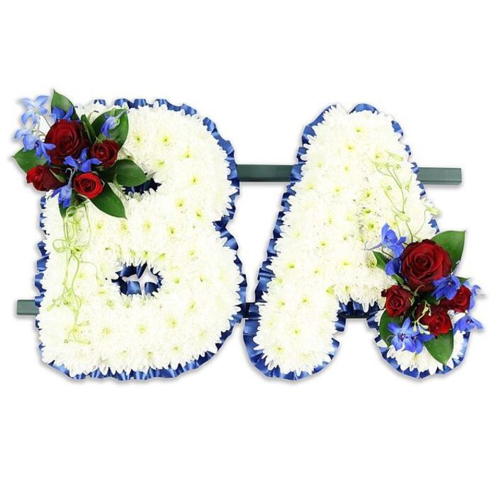 A dignified memorial composition shapes the capital letters BA in a paired, freestanding display, each letter densely packed with pristine white chrysanthemum blooms that form a soft, cloud-like plane of petals. The perimeter of both letters is carefully traced with a ruffled royal blue satin ribbon, the colour rich and somber, giving each form a defined silhouette that reads with clarity in a chapel or memorial setting. The B bears a floral accent on its upper-left curve: several deep red roses-their velvet petals moving from tight buds to gently unfurling blooms-nestle among spike-like, electric-blue delphinium-type flowers and deep green foliage, with a few pale green tendrils spilling softly to introduce movement. The A receives a matching cluster at its lower-right, mirroring the same red-and-blue motif so the two letters feel intimately connected. The dark green horizontal stand anchors the letters, assuring stability and unifying the composition; photographed against a stark white background, the colours appear particularly saturated-white reads as peace, blue as solemnity, and red as enduring love. There is a subtle floral perfume where chrysanthemum freshness meets the warmer, more familiar scent of roses; together these sensory notes reinforce the arrangement's purpose as a sympathy tribute. As a florist who understands local customs across Gerrards Cross, the Chalfonts, and nearby Iver Heath, I would recommend this style for a personalised funeral display, an inscription of initials placed beside a memorial photograph or altar as a final, visible message of respect and remembrance for family and friends.