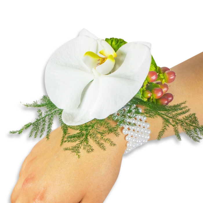 On a pale wrist against an immaculate white background sits a sophisticated floral corsage whose details are easy to read and cherish. Dominating the design is a single, large white Phalaenopsis orchid with broad, silky petals that curve open; its throat is painted in a bright yellow that deepens into a warm orange at the centre, creating a small, luminous focal point. The orchid rests on a bed of soft asparagus fern, its bright green, feathery fronds framing and cushioning the bloom while adding a sense of lightness and movement. To the upper right of the flower, a tight cluster of Hypericum berries offers a tactile counterpoint: each berry shows a subtle gradient from pale green through pinkish-rose to a rich cranberry, their glossy skins catching the light and suggesting freshness. A ruffled lime-green accent leaf nearby adds an extra layer of texture and contemporary colour. Everything is bound to the wrist with a multi-strand white pearl bracelet whose warm sheen and gentle weight lend an heirloom elegance to the piece. The overall effect is refined and romantic - a perfect accessory for a bride walking out from a Georgian townhouse on Gerrards Cross High Street or a prom-goer arriving by train at Gerrards Cross station. Crafted with local sensibility and an eye for classic detail, this corsage could be prepared by an experienced local studio such as Flower Delivery Gerrards Cross for special occasions across Gerrards Cross, Beaconsfield, and Chalfont St Giles.