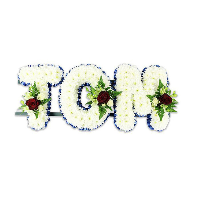 A bespoke sympathy piece that spells TOM in large, three-dimensional letters, hand-built from countless pure white chrysanthemums so densely placed they form a soft, tactile surface that seems to absorb light. Each letter's edge is precisely finished with a royal blue ribbon, a deliberate, crisp border that sets off the whiteness like a formal frame. Within the body of T, O and M sit matching floral sprays: a single deep red rose anchors each cluster, surrounded by small creamy spray roses and slender fern fronds that bring green veins of texture across the white expanse. The arrangement rests on a subtle dark green metal support at the base, engineered to stand at a funeral church or to be placed beside a graveside cross, and was photographed against a clean white background so the colours remain vivid and unequivocal. The scent profile is modest - a soft rose perfume threaded with fresh, leafy notes from the ferns - intended not to overwhelm but to offer calm. As a Gerrards Cross florist I see this composition as precisely the sort of tribute families request when arranging services along the High Street or for receptions close to the station or parish church: respectful, legible from a distance, and intimate up close. Flower Delivery Gerrards Cross creates pieces like this with attention to balance, proportion and the quiet symbolism of colour - white for solace, blue for dignity, and red for enduring love - fashioned to honour a life and to bring comfort to those gathered.