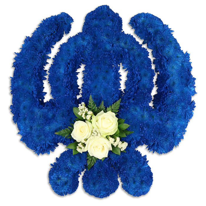 An intricately constructed Sikh Khanda floral tribute is displayed against a simple white backdrop, allowing the symbolic form and saturated colours to be fully appreciated. The florist has used countless royal blue chrysanthemum florets to map out the complete Khanda emblem: a strong vertical central sword, the enclosing circular chakkar, and the two elegant kirpans that curve outward like protective wings. Each tiny flower head is placed tightly beside the next, creating a solid field of rich blue that feels both powerful and serene, reminiscent of a ceremonial cloth or a beautifully woven tapestry. The edges of the Khanda are clean and well defined, reflecting careful planning and a steady, practiced hand. At the base of the central blade, a compact nosegay in white and green sits like a quiet prayer within the larger design. Three white roses, just at that perfect stage of opening where each petal can still be clearly seen, form the centrepiece of this cluster. Around them, fine sprays of white baby's breath add softness and light, while glossy green leaves introduce a fresh, natural contrast to the blue. Soft studio lighting highlights the texture of every petal and leaf, and gentle shadows give the emblem a slightly raised, almost sculptural presence. Designed as a meaningful funeral or memorial tribute for Sikh families in Gerrards Cross, from the tree-lined roads off Packhorse Road to homes near Bulstrode Park, this arrangement embodies respect, remembrance, and spiritual strength. It is the kind of floral piece an experienced local florist in Gerrards Cross would create when asked for something that honours both tradition and the unique life being remembered.