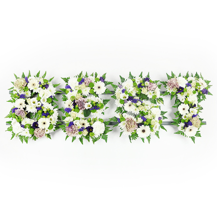 A meticulously composed, horizontal floral display spells BEST in four full, capital letters, set against a pristine white backdrop that highlights the fine details of colour and texture. Each letter is an abundant arrangement of predominantly white blooms-large, immaculate gerbera daisies with their dark centres, soft lisianthus, and rounded white carnations-layered to create a varied, tactile surface. Interspersed among these are subtle mauve and lavender roses and ruffled carnations that introduce dignified, subdued colour; tiny clusters of deeper purple filler flowers add shadowed depth and a quiet richness. The perimeter of every letter is softened and defined by feathery fern fronds, whose light, linear foliage frames the composition with organic grace. Within the floral fields, green button chrysanthemums and compact round greens punctuate the whites and purples, lending freshness and a pleasing contrast of form. A dark grey structural base underlies the letters and peeks through in places, providing the necessary definition that keeps each character legible while allowing the flowers to remain the visual focus. Under soft studio lighting the petals take on a gentle sheen and the whole tribute reads as serene and respectful-an arrangement crafted for memorial services, wakes, or cemetery placements. The scent is understated: green, clean, and floral rather than overpowering, suited to a moment of remembrance. Locally, this kind of personalised tribute is familiar to families across Gerrards Cross and nearby villages when they seek a composed, expressive floral message that honours a loved one with elegance and thoughtful floral design.