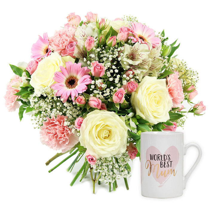 Studio image of a sentimental floral and mug gift set, styled with the understated elegance you might expect from a trusted Gerrards Cross florist. Against a clean white backdrop, a round, hand-tied bouquet dominates the left side, its stems carefully bound and visible at the bottom, hinting at the craftsmanship behind the design. At the centre are several large cream or pale yellow roses, fully open yet still structured, their petals layered in soft spirals that seem almost velvety to the touch. These classic blooms are framed by ruffled light pink carnations and clusters of darker pink spray roses, which give the arrangement a pleasing fullness and variation in size and shape. Bright pink gerbera daisies, each with a bold dark centre, punctuate the bouquet with a more contemporary feel, their flat petals radiating like little suns. Threaded through this pink-and-cream harmony are white alstroemeria, their slender petals etched with fine reddish-brown flecks that draw the eye on closer inspection. Wisps of baby's breath, with its tiny white flowers, sit like a delicate mist around the larger blooms, softening the whole composition. Touches of fresh green foliage peek out between the flowers, providing just enough contrast to keep the palette lively while still gentle. To the right stands a pristine white ceramic mug with a smooth, comfortable handle. On its front, a soft pink watercolour heart holds the words World's Best Mum, with World's Best printed in dark lettering above the word Mum, written in a shimmering gold or copper script that catches the light. The image suggests a heartfelt gesture, perfect for Mother's Day or a special birthday, conjuring the feeling of delivering something beautiful to a doorway near East Common or along the tree-lined streets of Gerrards Cross.