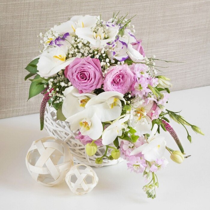This image shows a refined floral centrepiece styled in a round white wicker basket, as if prepared for a wedding table or elegant dinner party in Gerrards Cross. The basket is generously filled with a harmonious mix of pink and white flowers, creating a softly romantic palette. Front and centre, several pale pink roses are in full bloom, their petals curling into classic spirals that suggest both tradition and tenderness. These roses are framed by pristine white Phalaenopsis orchids whose broad, smooth petals form graceful arcs, each bloom revealing a warm yellow-orange centre that glows subtly against the white. Threaded among them are white Lisianthus flowers, some with a whisper of lavender-purple tracing their edges, lending a sense of delicate refinement. Stems of pale pink stock rise and then cascade gently over the rim, their many small blossoms adding vertical interest and a lightly textured backdrop for the more sculptural roses and orchids. Sprays of airy white Gypsophila appear throughout, scattering tiny blossoms like confetti and softening the overall outline. Deep and mid-green foliage nestles between the blooms, adding contrast and creating the impression of something freshly cut from a well-tended garden off Bulstrode Way or near West Common. The white wicker basket, tightly woven and neatly finished, sits on a bright white surface that reflects the natural light and lifts the colours of the flowers. To one side lie two decorative woven balls, crafted from pale, natural fibres that intertwine in open, spherical patterns mirroring the basket's rustic texture. The backdrop is a light, neutral fabric, gently textured and likely linen or similar, which keeps the focus squarely on the floral artistry. The entire composition conveys serenity, romance and thoughtful craftsmanship, making it perfect inspiration for wedding table arrangements or special occasion flowers delivered locally in Gerrards Cross.