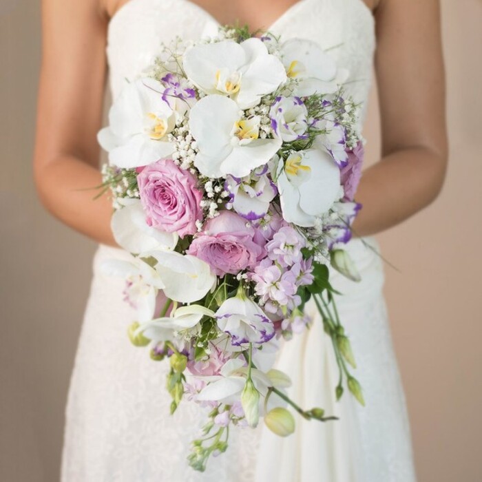 This radiant close-up captures a bride from the waist up in Gerrards Cross, elegantly holding a showstopping cascading bouquet that softly dominates the scene. Set against a smooth, blurred ivory backdrop, the bouquet unfurls downward in a fluid teardrop, blending pristine white orchids, lush garden roses in blush pink, and delicate lavender accents into a cohesive, luxurious composition. Each Phalaenopsis orchid commands attention with its broad, satiny petals edged in subtle yellow and hints of purple, their exquisite shapes often admired in local Buckinghamshire weddings. Amongst them, garden roses in various shades of pink and mauve cluster in romantic abundance, their plush, densely layered petals inviting both touch and admiration. Small, spiky lavender stock and intricate white lisianthus - some with whimsical purple-tipped petals - peek through, joined by wisps of white gypsophila that appear almost weightless. The bouquet's fresh green foliage is woven sparingly to add contrast without overwhelming the serene palette. The bride's simple yet refined strapless dress, its bodice detailed with intricate lace, subtly grounds the bouquet's exuberance, while her gently poised arms direct all focus to the meticulous arrangement. This image is a testament to the refined style and expert craftsmanship that characterise weddings in Gerrards Cross, making it an inspiring visual for anyone planning a heartfelt local celebration or seeking fresh bridal flower ideas firmly rooted in the beauty of the Chiltern Hills.