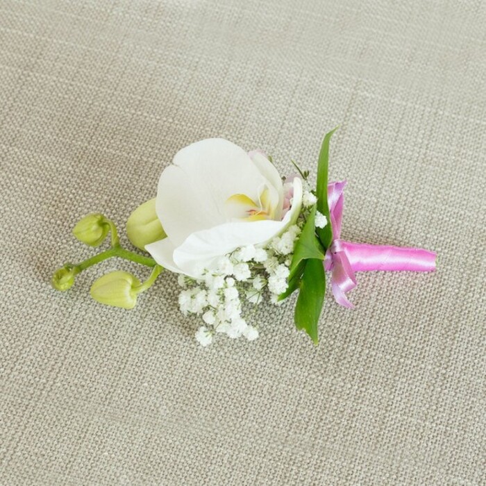 A single orchid boutonniere is displayed lying sideways on a neutral, oat-hued linen fabric, its gentle texture forming a quiet stage that allows every detail of the flowers to stand out with clarity. At the centre of the piece sits a pristine white Phalaenopsis orchid, its large, rounded petals elegantly arranged like folded silk. The bloom's heart reveals a refined blend of colour: a soft, buttery yellow and the subtlest hint of blush pink in the throat, adding depth to the otherwise pure white petals. Surrounding this central orchid, clusters of baby's breath create a light, cloud-like halo; their many tiny white blossoms and fine stems lend the accessory a soft, romantic air that would feel perfectly at home at a ceremony near the Gerrards Cross rail bridge or a reception overlooking the green spaces by East Common. A few slender, fresh green leaves appear behind the flowers, introducing a clean, natural contrast and a sense of movement. Extending toward one side, a short stem carries several unopened, lime-green orchid buds, tightly furled and promising future blossoms, which subtly balance the composition and contribute a feeling of gentle growth. The stems are gathered neatly and wrapped in a vivid fuchsia satin ribbon tied into a small, precise bow, the sheen of the fabric catching the light and providing a lively yet refined splash of colour. Soft, even lighting smooths across the scene, enhancing the velvety texture of the orchid petals, the airy volume of the baby's breath, and the fine weave of the linen. The overall impression is of a carefully crafted floral accessory, ideal as a groom's boutonniere or corsage for a special guest, thoughtfully designed by a local Gerrards Cross florist to add a touch of polished elegance.