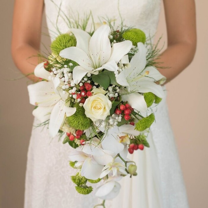 Capturing a moment just outside the grand churches and manor venues of Gerrards Cross, this image focuses on the spectacular beauty of a bridal bouquet, held just above the skirt of a softly cascading white lace gown. The luxurious arrangement showcases several prominent oriental lilies, each bloom pristine white with subtle cinnamon freckles painting their throats, the classic emblem of refined bridal taste. Nestled amidst the lilies is a perfectly formed cream rose, its folding petals tight at the center and whispering romantic promise. Sweeping gracefully downward from the bouquet's base, a trail of white phalaenopsis orchids creates a dynamic, cascading silhouette, giving the design its dramatic waterfall effect. Puff-like clusters of lime-green mums or pom-pom chrysanthemums are woven throughout, infusing the bouquet with a lively contemporary energy-much like the vibrant Gardens of Bulstrode Park nearby. Sprays of cloudlike white baby's breath soften the arrangement and offer an ethereal touch, while festive, glossy red berries, reminiscent of Gerrards Cross's beloved winter displays, punctuate the bouquet with warmth and jubilation. Wisps of green foliage and delicate, grass-like strands provide a natural framework, enveloping the bouquet in an organic aura. Every detail-from dew-kissed petals to ribbon finishing-reflects the careful hand of a local florist attuned to wedding trends and traditions in Gerrards Cross, making this bouquet a standout inspiration for any ceremony in the area.