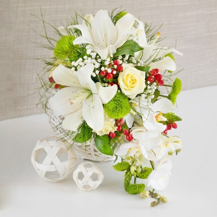 An exquisitely detailed table arrangement rests on a spotless white surface, the kind of refined floral design often chosen for wedding receptions and milestone celebrations around Gerrards Cross. The flowers are artfully gathered into a round, white woven basket whose lattice texture introduces a handcrafted, natural touch. Towering from the centre are several large white Oriental lilies, their trumpet-shaped blooms fully open, revealing subtle speckles and pollen-laden stamens that hint at a soft, sweet fragrance. Tucked just off-centre, a single cream-to-pale-ivory rose opens in layered perfection, its gentle warmth contrasting beautifully with the crisp white petals around it. From the right side of the arrangement, a delicate cascade of white Phalaenopsis orchids tumbles downward in a graceful curve, creating a striking waterfall effect often favoured in modern bridal designs. The composition is enriched with lush greenery: fine, airy foliage, slender grass-like strands, and broader, polished leaves that frame the flowers. Bright green spherical blooms, reminiscent of dianthus or green trick, nestle between the lilies, adding a unique, velvety texture. Scattered through the bouquet, tiny white baby's breath blossoms soften transitions, while clusters of deep red berries punctuate the whites and greens with jewel-bright colour, evoking the hedgerows that line the lanes approaching Bulstrode Park. To the left of the main display, two white woven spheres of different sizes rest calmly on the table, echoing the basket's material and adding sculptural interest to the scene. Behind it all, a softly textured beige backdrop, likely linen or a similar fabric, diffuses the light, ensuring every petal, leaf, and berry is gently defined. The arrangement conveys a sense of purity, celebration, and understated luxury, the sort of piece that could anchor a wedding table or formal gathering anywhere in and around Gerrards Cross.