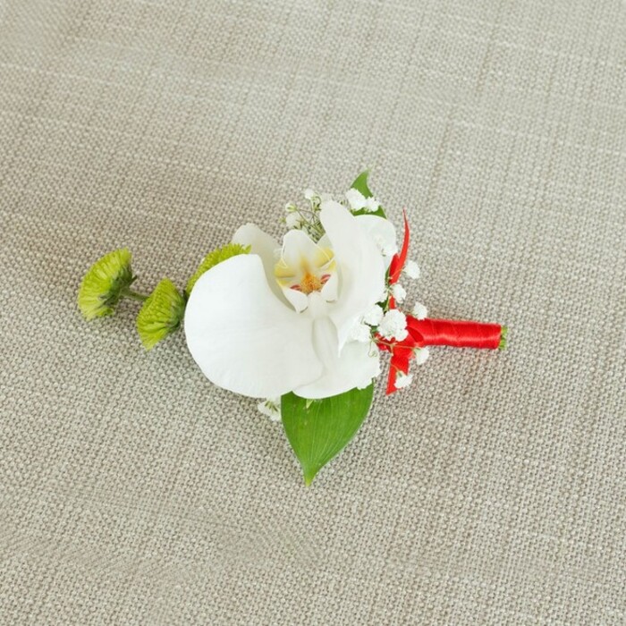 On a softly lit, light beige linen background, a single, exquisitely composed boutonniere is presented as though waiting to be pinned to a jacket just before a ceremony in Gerrards Cross. The fabric beneath it has a visible, slightly coarse weave, suggesting natural fibres that lend a calm, organic feel and allow every floral detail to stand out. At the heart of the piece is a large white Phalaenopsis orchid, its petals broad, gently curved, and satin-smooth, folding around a central column patterned with soft yellow and delicate reddish-orange striations. These inner markings add a quiet complexity, like fine brushstrokes on a porcelain surface. Encircling the orchid, clusters of baby's breath spill outward, each tiny white blossom a dot of light, forming an ethereal halo that softens the edges of the design. To the left of the orchid, two lime-green chrysanthemum-style pom-pom buds introduce a lively contrast; their compact, spiky petals seem almost moss-like, bringing a fresh, modern twist to the otherwise classic pairing of white and green. A strong, deep green leaf supports the composition from below, with a smaller leaf nestled near the base, giving the arrangement structure and a sense of natural growth. The stems are bound with a vivid crimson red satin ribbon, wrapped smoothly and securely, finishing in a small, elegant knot that adds a note of celebration reminiscent of ribbon used for special events across Buckinghamshire. The overall impression is of thoughtful craftsmanship and quiet luxury, perfect for a groom, best man, or guest attending a local wedding, perhaps in one of the refined venues around East Common, where understated details make lasting memories.