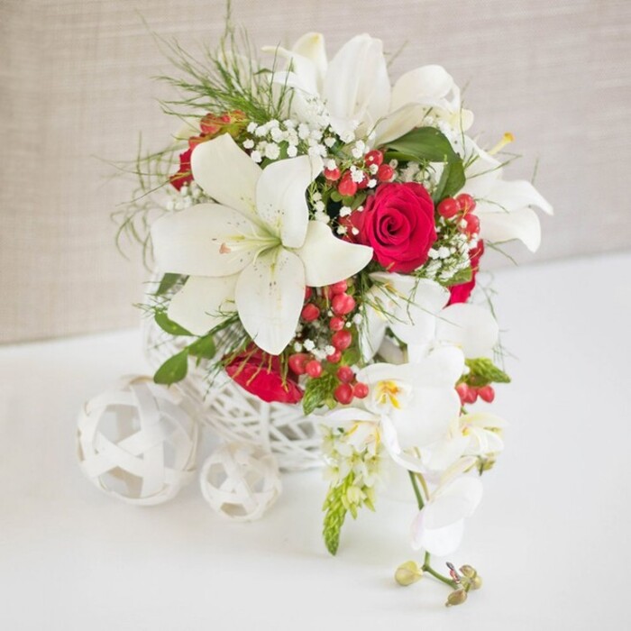 A luxurious floral arrangement, designed as an elegant wedding or celebration centerpiece, rests on a smooth white surface that subtly reflects its colours, as if set on a polished table in an airy Gerrards Cross dining room. The backdrop is a soft, light beige textured fabric that provides a warm, neutral canvas, allowing every petal and leaf to stand out clearly. At the heart of the arrangement is a round, white woven container with an open, basket-like pattern, its delicate latticework echoing the look of hand-crafted rattan and adding a relaxed yet refined feel. Rising from this base, several large white oriental lilies command attention, their six broad petals elegantly curved and slightly ruffled at the edges, with long, slender stamens tipped in golden-brown pollen. Between and around these lilies, deep red roses appear like jewels, their petals densely layered and velvety, suggesting passion and enduring love. Fine sprays of white gypsophila, or baby's breath, drift through the composition, softening the strong shapes and creating a gentle, cloud-like haze. Bright green wisps of foliage, airy and feathery, add movement and a sense of freshness, reminiscent of the greenery along Bulstrode Way on a spring morning. Scattered among the blooms, glossy red hypericum berries catch the light, bringing subtle festive energy. On one side, an elegant cascade of white Phalaenopsis orchids spills down from the arrangement, each bloom with porcelain-like petals and a delicate yellow and blush centre, accompanied by a few tight buds that promise future beauty. To the left of the main display, a trio of white woven spheres-each slightly different in size-echoes the container's material, adding sculptural interest. The completed design feels opulent yet tasteful, the kind of piece Flower Delivery Gerrards Cross might place for a wedding breakfast, anniversary dinner, or sophisticated celebration in one of the town's charming venues.