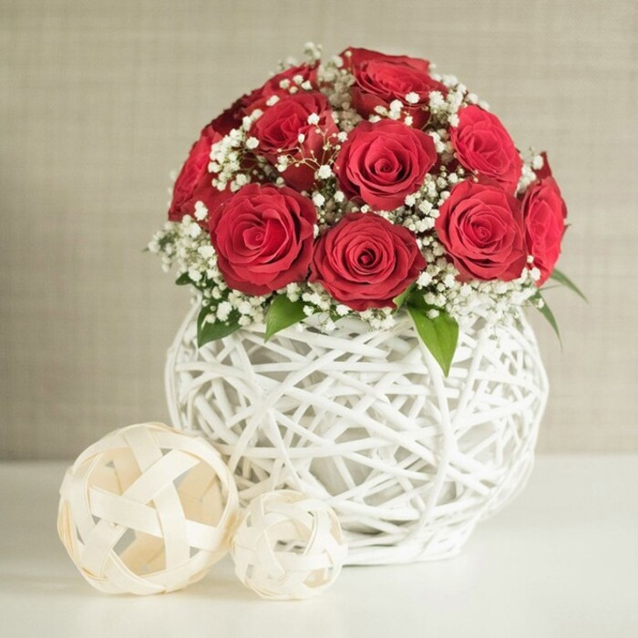 An elegantly styled floral centrepiece commands attention, designed in the kind of timeless palette that suits a wedding table in Gerrards Cross or a sophisticated dinner party at home. The arrangement is dominated by a rounded mass of deep red roses, arranged closely together so that each blossom brushes gently against the next, creating a continuous dome of colour. Every rose is at its prime, petals unfurled in spirals that transition from a tightly coiled centre to softer, more open edges, giving the bouquet a plush, almost velvety texture. Between the roses, soft sprays of white baby's breath nestle delicately, their tiny blossoms floating like light mist against the intense red. A few discreet green leaves emerge at the base and between the blooms, adding a whisper of natural contrast and hinting at the garden origins of the flowers. This luxurious cluster is set in a distinctive, spherical white vase whose structure is formed from tangled, interwoven strands that criss-cross and loop, leaving glimpses of the stems within. The vase has an airy, lattice-like quality, reminiscent of modern woven décor pieces often seen in stylish homes around the Chilterns. On the polished white surface in front and slightly to the side of the vase rest two decorative balls in an off-white or creamy hue, also created from slender strips woven into open, criss-cross patterns. One sphere is noticeably larger, anchoring the composition, while the smaller one is tucked behind, lending a pleasing sense of scale. The backdrop is a soft, neutral wall, subtly textured and softly lit, which keeps the atmosphere calm and intimate. The overall mood is one of refined romance and quiet celebration, ideal for capturing the feel of a wedding breakfast or engagement dinner in and around Gerrards Cross.