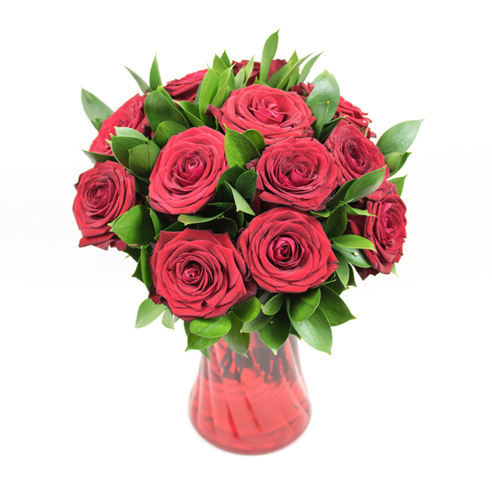 In this image, a rounded bouquet of approximately twelve to fifteen deep crimson roses is showcased in a translucent red glass vase, positioned against a spotless white background that allows the colours to stand vividly on their own. The roses form a tight, almost perfectly spherical dome, each bloom fully open yet still crisp, with layers of velvety petals swirling inward in elegant spirals. Their colour is a saturated ruby red, intense and full-bodied, suggestive of classic romance and carefully selected quality. Nestled among the blooms are generous sprigs of dark green foliage, likely ruscus or a similar leafy green, its smooth, pointed leaves providing a lush, supportive framework. The greenery slips between petals and peeks out around the edges, giving the arrangement volume and a natural, garden-like character rather than a rigidly formal look. The vase is fashioned from rich red glass with a gently flared neck and a softly curving body that narrows as it reaches the base, offering both visual grace and sturdy support. Subtle ribbing or swirling along the surface catches and reflects light, creating delicate variations in tone that mirror the complexity of the rose petals above. The stems are faintly visible through the tinted glass, adding a sense of authenticity and craftsmanship. Against the bright white studio setting, every shadow is soft, every highlight precise, emphasizing the bouquet as a singular focal point. The overall mood is one of timeless devotion, an arrangement that could easily be imagined arriving from Flower Delivery Gerrards Cross to a home near West Common, ready to mark a milestone anniversary, a Valentine's gesture, or a special evening where love and appreciation take centre stage.