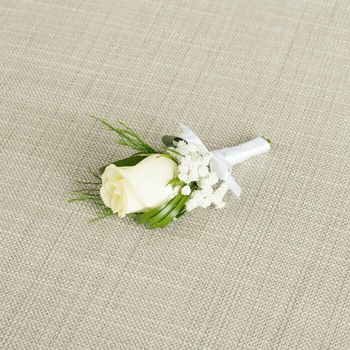 An elegant white rose boutonniere rests at a gentle angle on a light beige textile that resembles natural linen, its visible crosshatch weave adding a touch of rustic refinement. The focal point is a single creamy white rosebud, tightly but not rigidly furled, its outer petals curving in soft, smooth layers that hint at a full bloom yet to unfurl. Around this central rose, tiny florets of white baby's breath cluster like delicate stars, their fine stems and miniature blossoms giving the arrangement a soft, ethereal halo. Tucked behind and alongside the rose are elongated, dark green leaves and a few more feathery sprigs of foliage, their shapes overlapping in a way that frames the bloom and creates a subtle sense of movement. The stems of the rose and greenery have been expertly bound with a band of white satin ribbon, wound snugly and finished with a small, precise bow positioned just below the flower head, clearly indicating that this piece is ready to be pinned onto a smart jacket. The lighting is diffused and gentle, casting soft shadows that bring out the curve of each petal, the airy texture of the gypsophila, and the faint sheen of the satin. The whole scene conveys the calm anticipation of a wedding morning in Gerrards Cross, perhaps before a ceremony at a local church or a celebration in a nearby country venue. Crafted with the care and precision you would expect from a neighbourhood florist in the area, this timeless white boutonniere would suit a groom, best man, or usher arriving from nearby streets like Fulmer Road or Austenwood Lane, capturing the purity, new beginnings, and understated luxury that define many local celebrations.
