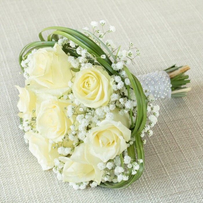 Presented against a backdrop of subtle beige linen, this stunning bridal bouquet from a Gerrards Cross florist is the epitome of refined artistry and classic romance. Seven or eight creamy white roses, captured in various luminous states of bloom, create an unmistakable focal point. Their delicate petals form gentle spirals, inviting a closer look at the careful arrangement. Fluffy clusters of baby's breath (Gypsophila) nestle among the roses like miniature clouds, imparting a dreamy softness and graceful fullness to the bouquet-evoking the tranquil atmosphere of a stroll along West Common. Threaded throughout are slender loops of verdant bear grass, artfully arranged to add movement and fresh energy while highlighting the bouquet's sophisticated structure. Each stem is gathered and expertly wrapped in a shimmering silver ribbon, its length studded with neat parallel rows of lustrous white pearls, providing a luxurious tactile detail. Below the ribbon, the natural cut stems peek through, reinforcing the bouquet's hand-tied authenticity. Designed for celebrations large or small-whether a garden wedding on Oxford Road or an intimate anniversary gathering-this bouquet beautifully captures timeless elegance and the spirit of special occasions in Gerrards Cross.