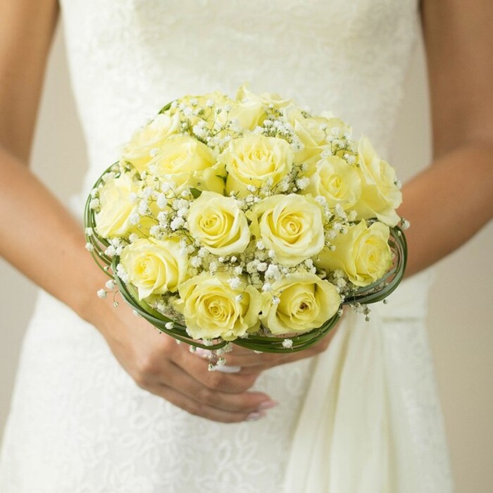 This evocative scene from a Gerrards Cross wedding offers a close-up perspective of a bridal bouquet, held with heartfelt care by a bride in a delicately textured white gown. Central to the image is the bouquet-a perfectly rounded gathering of pale yellow roses, each blossom plush and at the peak of bloom, their centers unfolding in a gentle swirl. Wispy tendrils of white baby's breath weave among the roses, providing a cloudlike, almost dreamlike contrast that elevates the entire composition. Surrounding the flowers at the base are slender, dark green leaves or intricately coiled decorative wire that add sophistication and subtly reference the rich greenery of Gerrards Cross's local parks. The bride's hands-fingers adorned with a simple wedding band and nails glossed in a pale neutral hue-rest beneath the bouquet, cradling it as if treasuring both the tradition and the fresh promise it represents. The soft blur of the background, punctuated by hints of lace or embroidery, accentuates the rich chromatic interplay between white, yellow, and green, while creating an atmosphere of gentle celebration. This arrangement, thoughtfully designed for a Gerrards Cross ceremony, captures the grace, romance, and meticulous floral artistry that brides from the area cherish for their wedding day.