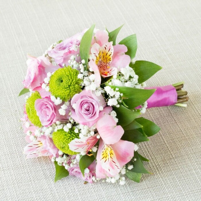 Artfully arranged by a skilled florist in Gerrards Cross, this elegant bouquet radiates charm and freshness as it sits diagonally on a subtly textured, pale beige fabric, reminiscent of the local market's understated elegance. Anchoring the arrangement are several pastel pink roses, their gently swirling petals unfolding like stories shared between friends at a Gerrards Cross café. Nestled amid the roses, vibrant lime-green button chrysanthemums add bursts of playful energy, while delicate sprays of pink alstroemeria open to reveal streaks of golden yellow and intricate burgundy freckles. These touches evoke the graceful wildflowers seen on woodland walks near Bulstrode Park. Soft, cloudlike clusters of baby's breath (gypsophila) fill the spaces between blooms, infusing the bouquet with a dreamlike softness and unity. Rich green foliage envelops the flowers, enhancing their hues and offering a lush, layered depth akin to the area's verdant gardens. At the bouquet's base, a glossy magenta satin ribbon is wrapped carefully around the stems, binding them into a harmonious whole and adding a sophisticated finish-ideal for a bridesmaid to carry down the aisle, or as a heartfelt gift delivered to a loved one in Gerrards Cross. Bathed in gentle, diffused lighting, every petal's subtle sheen and dewy texture is caught to perfection, making this arrangement a radiant testament to thoughtful floral artistry and local celebration.