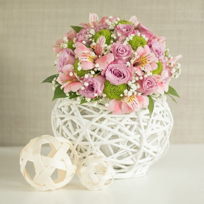 A sophisticated floral centerpiece commands attention on a white tabletop, celebrating both classic romance and modern artisan style. The bouquet's abundant pastel pink and lavender roses evoke the genteel spirit of Gerrards Cross, harmoniously paired with pale pink alstroemeria-each bloom marked by delicate inner stripes suggestive of the subtleties seen in nature along the River Misbourne. Scattered among the larger flowers, spheres of lime green button chrysanthemums offer vibrant, textural contrast, while accents of pure white baby's breath weave between the blooms, imparting a sense of airiness akin to springtime fresh air at Gold Hill Common. The striking white vase, crafted in an open, lattice weave from slender white strips, stands as a statement in contemporary rustic design, artfully blending sophistication with homey warmth cherished in the local community. Two decorative, woven spheres in soft cream and beige tones rest nearby; their sizes vary, enhancing visual interest and balancing the composition in a nod to Gerrards Cross's unique blend of heritage and modern vibrancy. The whole scene, bathed in gentle natural light with a soft textured beige backdrop, radiates calm and joyful celebration-perfect as a wedding centerpiece or an elegant statement for local special events. Every detail, from petal to weave, reflects the refined care of skilled local florists.