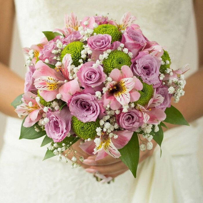 Held gracefully against the sumptuous texture of a white wedding gown, this bridal bouquet embodies the artistry and fresh elegance beloved in Gerrards Cross weddings. Central to the arrangement, clusters of blush pink and lavender roses bloom at multiple stages, some petals still adorned with morning dew. Weaving between these romantic classics, lime-hued button chrysanthemums pop with joyful energy, their soft, round forms adding depth and a modern zest to the overall design. Pink alstroemeria lilies, marked with filigreed maroon and golden streaks, are artfully tucked in, bringing an unexpected touch that hints at far-flung gardens beyond the Chilterns. Ethereal white baby's breath clusters knit the composition together, lending an airy, dream-like quality evocative of spring mornings in Gerrards Cross or a stroll through West Common's meadows. The deep green leaves ground the palette, heightening each flower's vividness and forming a harmonious frame. In the bride's gentle grasp, the bouquet feels deeply personal-an expression of love and a fresh beginning, hand-crafted for special ceremonies held at local churches or country manors. This visual narrative celebrates not only the flowers but also the expertise of local florists, capturing the spirit of joyous unions in this peaceful Buckinghamshire community.