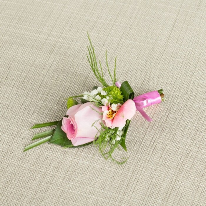 Diagonally placed atop a gently textured, cream-toned linen, this meticulously arranged boutonniere reflects the floral artistry found in the heart of Gerrards Cross. The spotlight shines on a single, flawless pink rosebud-its tender petals elegantly coiling from a deep rose core to delicately pale tips, capturing the fresh charm seen in spring gardens along Packhorse Road. Nestled close is a beautiful alstroemeria in shades of blush and peach, quietly unfurling petal by petal, its center highlighted by subtle yellow tones and delicate dark lines-a nod to the intricate wildflowers that flourish in places like Austenwood. Accentuating these focal blooms are clusters of pure white baby's breath, softly cloud-like, and pearl-sized green button chrysanthemums, their zesty hue and pillowy texture infusing vitality. Surrounding foliage is layered for shape and contrast: airy, grass-like shoots and feathery fern tendrils arc upwards, while deeper green leaves at the base anchor the compact arrangement and gently cup the blooms. A carefully wrapped broad leaf at the stem's top provides added structure, and the stems are elegantly gathered with a vivid fuchsia satin ribbon, tied in a petite bow that radiates celebration, much like the cheerful gatherings at local venues. Illuminated by soft, diffused daylight, every petal and leaf is brought to life, their colors and textures standing proudly against the oatmeal fabric below. Perfect for Gerrards Cross weddings, proms, or formal events, this boutonniere is a refined floral statement, rich in local character and crafted with keen attention to detail.